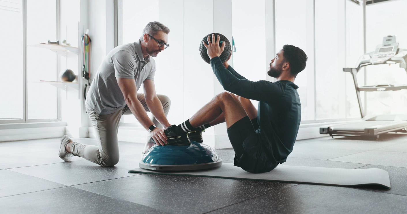 A physical therapist guiding a male athlete during an injury rehabilitation exercise. The athlete is seated on a balance trainer while holding a medicine ball, performing core-strengthening movements. The setting is a bright, modern gym with fitness equipment in the background